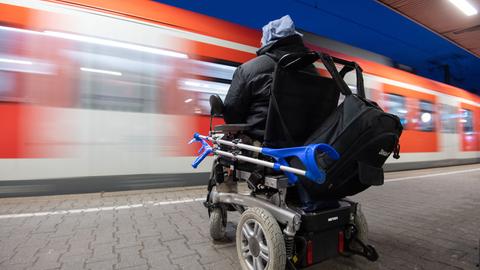 Ein Mann im Elektrorollstuhl sitzt an einem Bahngleis, ein Zug fährt an ihm vorbei