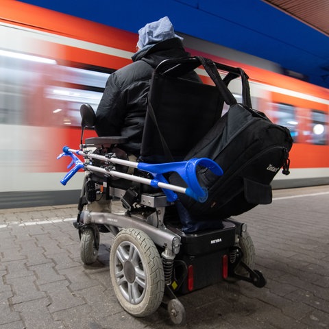 Ein Mann im Elektrorollstuhl sitzt an einem Bahngleis, ein Zug fährt an ihm vorbei