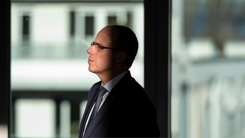 Innenminister Peter Beuth in einem Bürogebäude aus der Ferne fotografiert. Durch das Licht-Schatten-Spiel liegt sein Hinterkopf im Dunkeln und sein Gesicht im Licht. Er schaut nachdenklich nach links.