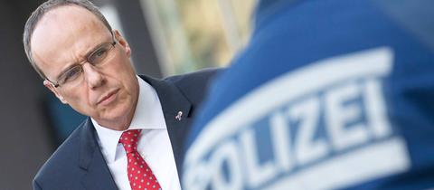 Innenminister Peter Beuth. So fotografiert, dass er auf den Rücken eines Polizeibeamten zu schauen scheint, auf dessen Rücken "Polizei" steht.