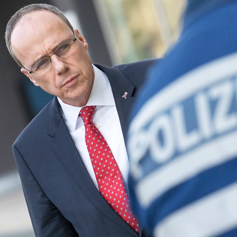 Innenminister Peter Beuth. So fotografiert, dass er auf den Rücken eines Polizeibeamten zu schauen scheint, auf dessen Rücken "Polizei" steht.