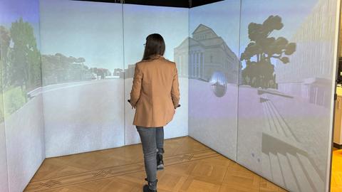 Eine Frau steht in einem kleinen abgetrennten Bereich, auf dem drei Beamer den Schlossplatz von Darmstadt in einer digitalisierten Variante projizieren. 
