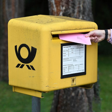 Hand, die einen Brief in einen gelben Briefkasten steckt. Rechts unten ein Wahlkreuz