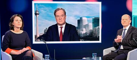 Annalena Baerbock und Olaf Scholz sitzen im Fernsehstudio in Sesseln, daneben kleine Tische mit Wassergläsern, bei einer Diskussionsrunde. Armin Laschet wird auf einem Fernsehschirm gezeigt.