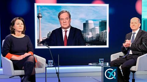 Annalena Baerbock und Olaf Scholz sitzen im Fernsehstudio in Sesseln, daneben kleine Tische mit Wassergläsern, bei einer Diskussionsrunde. Armin Laschet wird auf einem Fernsehschirm gezeigt.