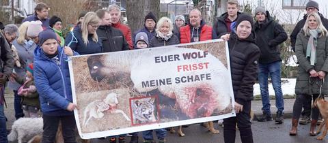 Menschen stehen auf einer Straße in einer Gruppe zusammen und halten ein Transparent hoch auf dem steht: "Euer Wolf frisst meine Schafe"