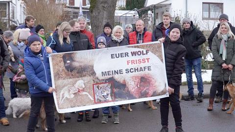 Menschen stehen auf einer Straße in einer Gruppe zusammen und halten ein Transparent hoch auf dem steht: "Euer Wolf frisst meine Schafe"