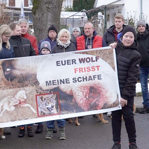 Menschen stehen auf einer Straße in einer Gruppe zusammen und halten ein Transparent hoch auf dem steht: "Euer Wolf frisst meine Schafe"