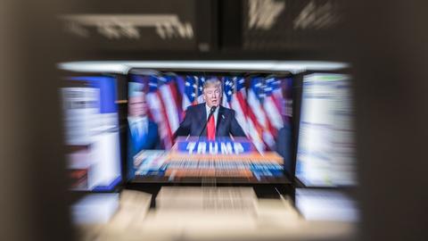 US-Präsident Trump auf einem Bildschirm, umgeben von weiteren Bildschirmen, auf denen verschwommen DAX-Kurse zu erkennen sind