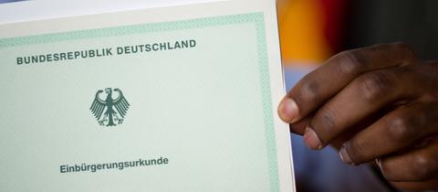 Eine Hand hält ein grünes Papier hoch, auf welchem "Einbürgerungsurkunde" steht.