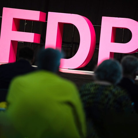 Pinkfarbene Buchstaben "F", "D", "P" stehen beleuchtet auf einer Erhöhung. Davor sind die Rücken von Menschen, die im Dunkeln sitzen, zu sehen.