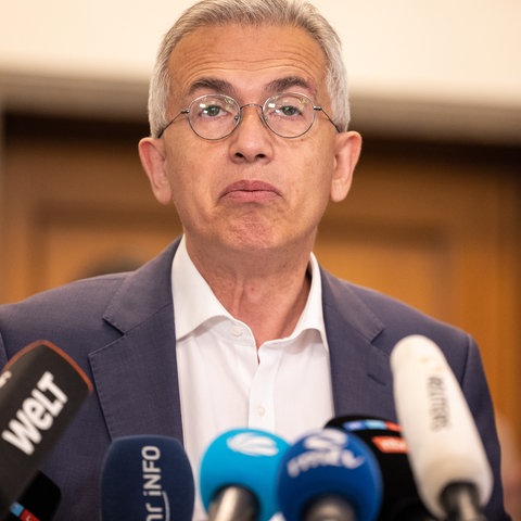 Peter Feldmann steht an einem Pult mit Mikrofonen und schaut auf - dabei mit fragendem Blick die Mundwinkel nach unten ziehend.