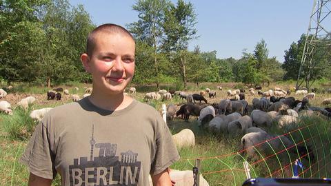 Eine junge Frau lächelt in die Kamera. Hinter ihr ist eine Schafherde auf einer Wiese zu sehen.