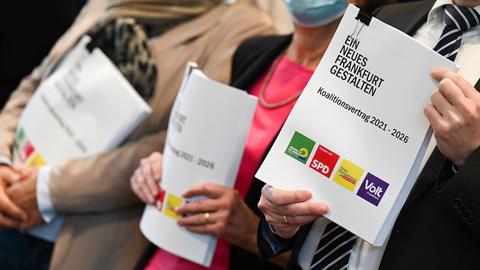 Drei Menschen stehen nebeneinander und halten ein dickes Schriftstück mit dem Titel "Ein neues Frankfurt gestalten" in der Hand. Am unteren Ende sind darauf die Logos der Parteien DIe Grünen, SPD, FDP und Volt zu sehen.