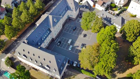 Zu sehen ist eine Luftaufnahme des Rathauses Friedberg. Wir schauen herunter auf ein weiß-braunes, herrschaftliches Gebäude mit einem schwarzen Dach. Davor ein leerer Betonparkplatz, drumherum Wohnhäuser und grüne Bäume