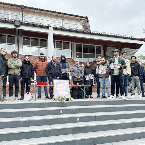 Menschen stehen an einer Treppe in einer Reihe zusammen. Vor ihnen ein Tisch mit Kerzen, Blumen und ein Foto eines Mannes.