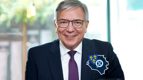 Mann mit grauen Haaren lacht in die Kamera. Auf dem Bild ein kleines "Label", bestehend aus einem Wahlkreuz, dem Wappen von Wiesbaden und dem Umriss der Stadt.