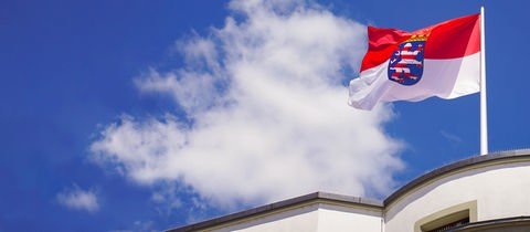 Im Bildvordergrund eine hessische Fahne, die auf dem Dach eines Gebäudes steht. Sie weht im Wind vor blauem Himmel mit Wolke.