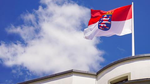 Im Bildvordergrund eine hessische Fahne, die auf dem Dach eines Gebäudes steht. Sie weht im Wind vor blauem Himmel mit Wolke.