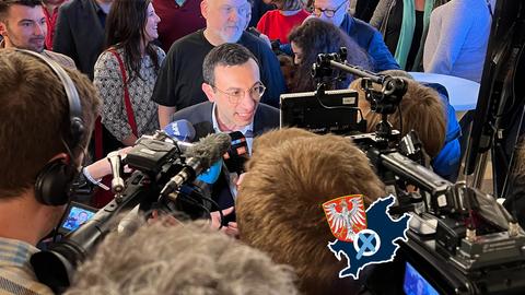 Foto von Mike Josef, umringt von Presse. Auf dem Foto eine kleine Grafik mit einer blau eingefärbten Fläche (Umriss Stadt Frankfurt), dem Wappen der Stadt Frankfurt und einem Wahlkreuz. ()