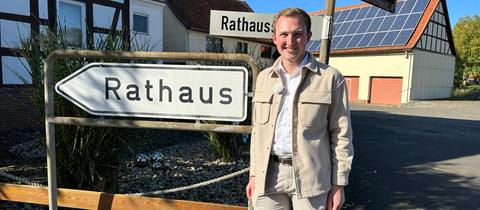 Lukas Becker, der neugewählte junge Bürgermeister von Lautertal, steht in Hemd und beiger Jacke neben einem Schild, auf dem Rathaus steht