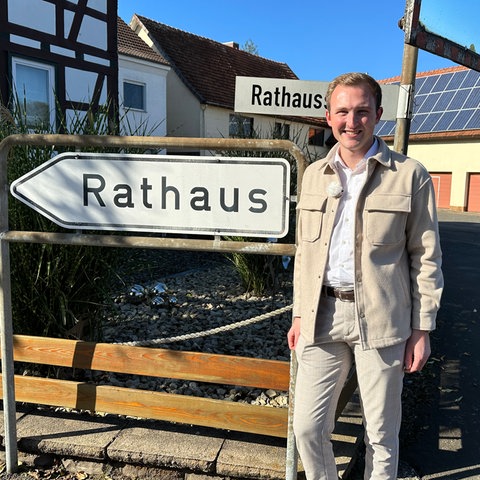 Lukas Becker, der neugewählte junge Bürgermeister von Lautertal, steht in Hemd und beiger Jacke neben einem Schild, auf dem Rathaus steht