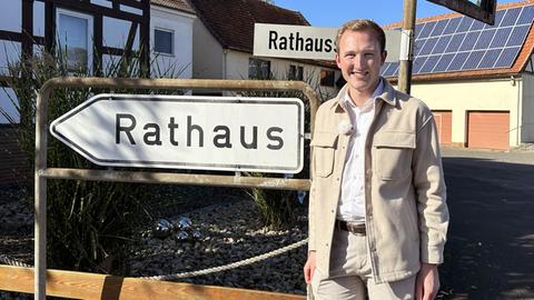 Ein jungenr Mann steht in Hemd und beiger Jacke neben einem Schild, auf dem "Rathaus" steht