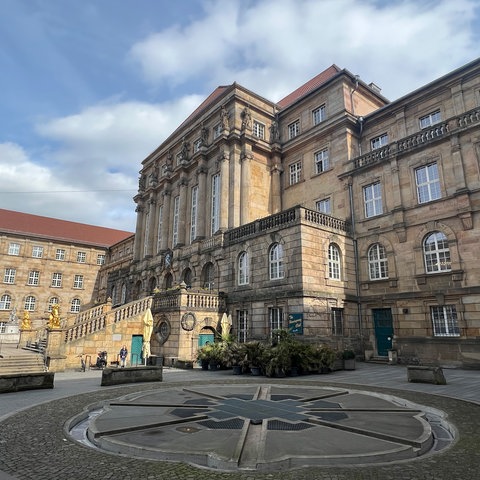 Das Rathaus in Kassel, zu sehen ist die große Treppe aus Stein, rechts und links zwei goldene Löwen. Im Vordergrund ist der Aschrottbrunnen zu sehen.