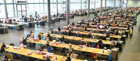 Unzählige Wahlhelfer*innen sitzen an sehr vielen Tischen in der riesigen Messehalle und zählen die Briefwahlstimmen aus (aus der Vogelperspektive fotografiert). 