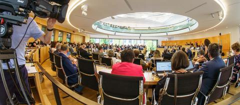 Fotot einer Plenarsitzung im hessischen Landtag. Im Vordergrund ein Kameramann an der Seite des Bildes.