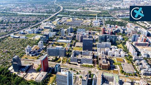 Luftbild eines Gewerbegebiets mit Hochhäusern, links davon eine Autobahn, im Hintergrund ein Fluss. Rechts oben ein Wahlkreuz.