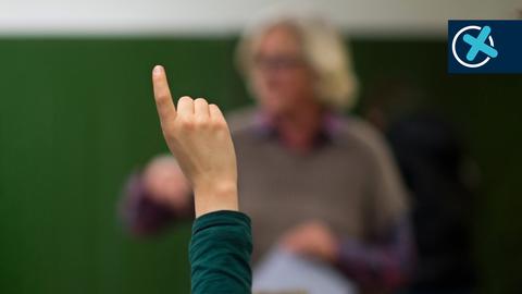 Schülerarm mit Finger nach oben im Bildvordergrund scharf. Im Bildhintergrund unscharf ein Lehrer vor einer Tafel stehend. Auf dem Bild rechts oben ein Wahlkreuz