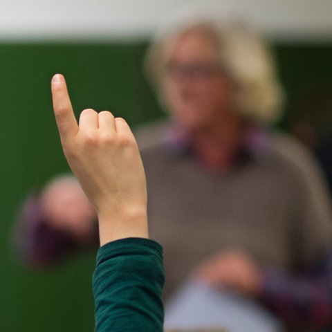 Schülerarm mit Finger nach oben im Bildvordergrund scharf. Im Bildhintergrund unscharf ein Lehrer vor einer Tafel stehend. Auf dem Bild rechts oben ein Wahlkreuz