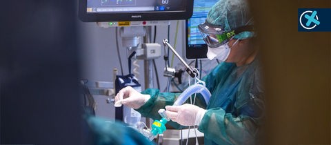 Foto: Blick durch eine Tür. Eine Intensivpflegerin in Schutzkleidung mit Schutzmaske und -brille arbeitet an einem Patienten, der mit Schläuchen und Monitoren verbunden ist.