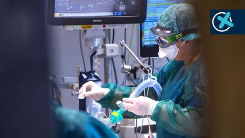 Foto: Blick durch eine Tür. Eine Intensivpflegerin in Schutzkleidung mit Schutzmaske und -brille arbeitet an einem Patienten, der mit Schläuchen und Monitoren verbunden ist.