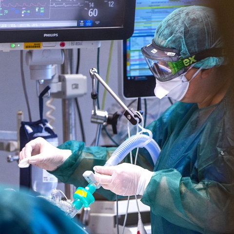 Foto: Blick durch eine Tür. Eine Intensivpflegerin in Schutzkleidung mit Schutzmaske und -brille arbeitet an einem Patienten, der mit Schläuchen und Monitoren verbunden ist.