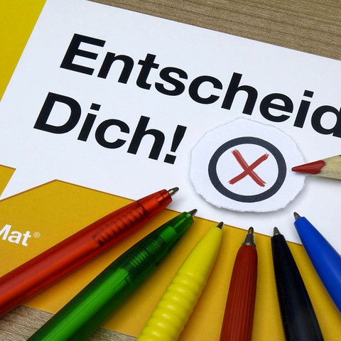 Zettel mit Aufschrift "Entscheide dich! Wahl-o-mat" und Stiften daneben in verschiedenen Farben, die Parteien zugeordnet werden können (blau, rot, grün, geld, usw.)