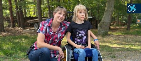Silke Hable hat den Arm um ein im Rollstuhl sitzendes Mädchen gelegt. Beide lächeln.