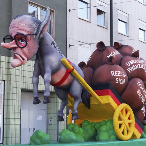 Ein KARNEVALSWAGEN BEIM ROSENMONTAGSZUG ， ZIEGT ALS PAPPMASCHEEFIGUR CDU-CHEF-CHEF-CHEF-CHEF MERZ ALS ESEL。 Er Zieht Einen Karren Mitsäcken, Uner Anderem Zum Schuldenpaket.