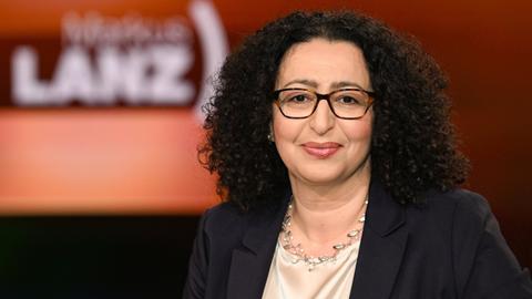 Portrait einer Frau. Im Hintergrund unscharf eine TV-Studio mit Aufschrift der Sendung "Markus Lanz".