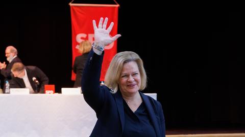 Nancy Faeser (SPD) winkt strahlend den Delegierten einer SPD-Versammlung zu, im Hintergrund drei Delegierte und eine SPD-Fahne