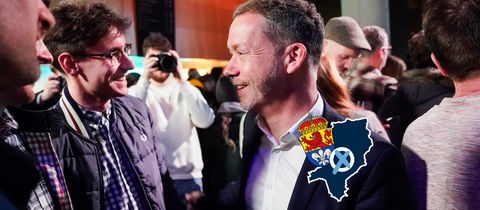 Foto von Kolmer, im Gespräch mit Menschen. Auf dem Bild eine kleine Grafik mit einer blau eingefärbten Fläche (Umriss der Stadt), dem Wappen der Stadt Darmstadt und einem Wahlkreuz.