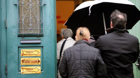 Ein Mann und eine Frau gehen mit einem Schirm vorm Regen geschützt zu einer alten, grünen Eingangstür. Darauf kleben Hinweisschilder mit der Aufschrift "Wahllokal". 