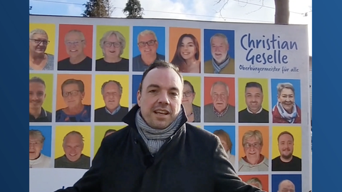 Christian Geselle, OB-Kandidat für Kassel, vor einem Werbeplakat mit Gesichtern von Bürgern