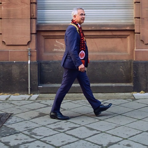 Peter Feldmann im Anzug, mit einem Eintracht-Schal um den Hals, geht auf einem Bürgersteig. Im Hintergrund herabgelassene Rolläden im Hochparterre eines historischen Hauses.