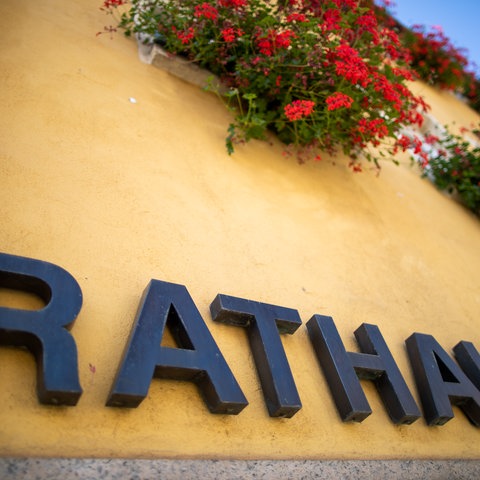 Schriftzug "Rathaus" an einer gelb gestrichenen Hauswand neben einer Blumenampel.