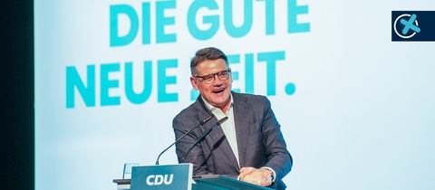 Ein Mann steht an einem Pult und spricht in ein Mikrofon. Am Pult steht "CDU". Im Hintergrund eine große türkisfarbene Wand auf welcher "Auf in die gute neue Zeit" in großen Lettern geschrieben steht.