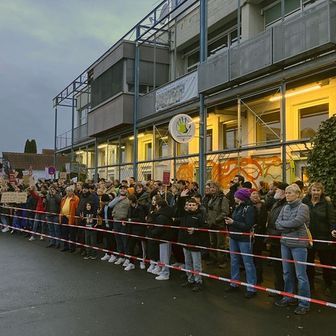 Dutzende Menschen stehen an einer mit Flatterband abgesperrten Straße und halten Plakate hoch. 