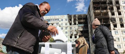 Eine Frau wirft einen Wahlzettel in eine Wahlurne, die auf der Straße vor zerstörten Gebäuden steht.