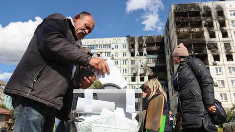 Eine Frau wirft einen Wahlzettel in eine Wahlurne, die auf der Straße vor zerstörten Gebäuden steht.